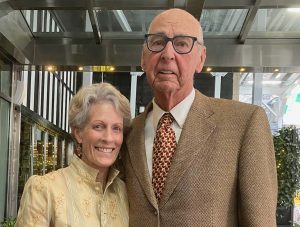 An older woman and man stand side by side, smiling with warmth. Proud Bend locals with a big heart, they pose outdoors near a glass building—the woman in a patterned jacket, the man in glasses and a brown suit with a patterned tie.
